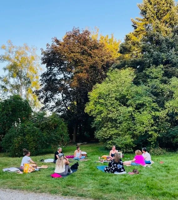 Eine Gruppe von Frauen sitzt im Kreis auf der Wiese vor einem Baum und hört einer Sprecherin zu.