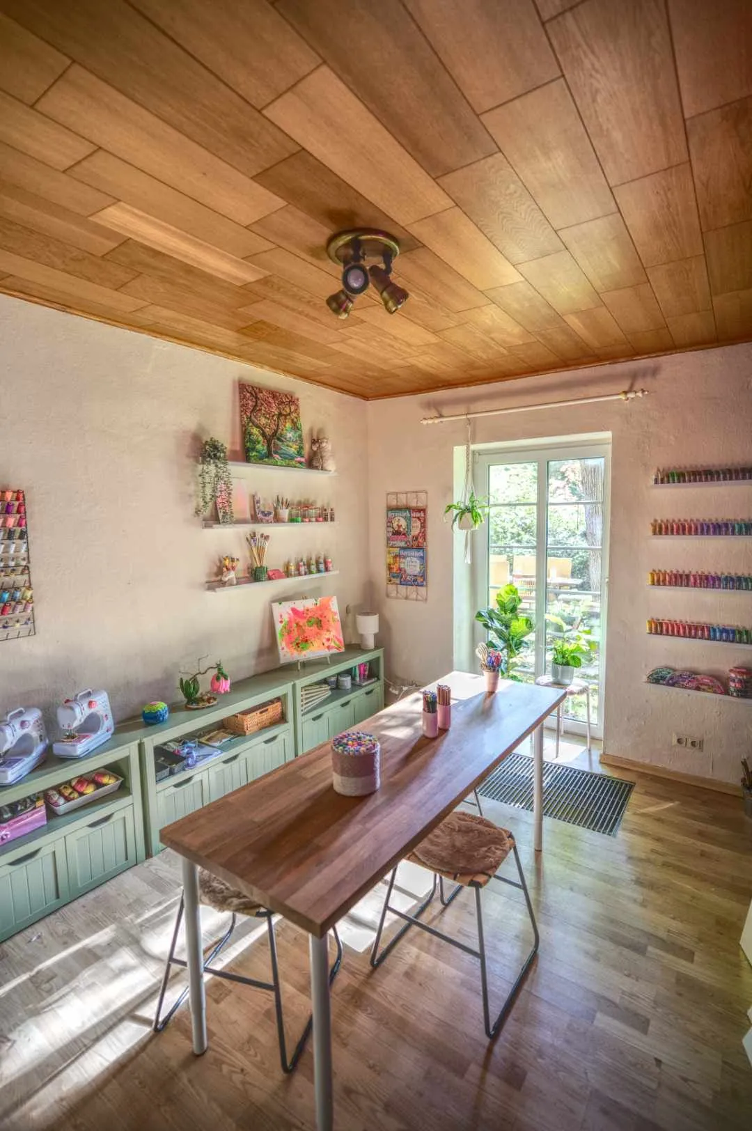 Ein helles Bastelzimmer mit einer hölzernen Decke, einem Holztisch mit Stühlen, mehreren Regalen mit Bastelmaterialien und einem großen Fenster mit Blick nach draußen.
